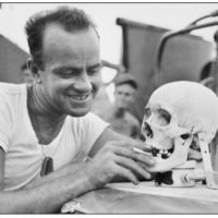 An American serviceman offers a cigarette to the skull of a Japanese soldier